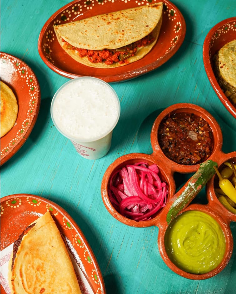Mesa servida con gorditas, tacos, salsas, cebolla morada y agua de horchata